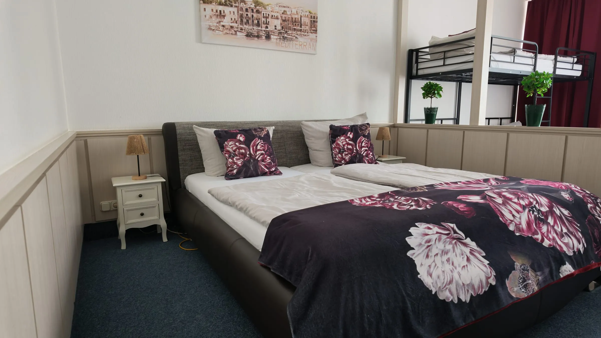 Doppelbett mit Blumendecke vor heller Holzvertäfelung, dahinter durch einen Raumteiler das Hochbett