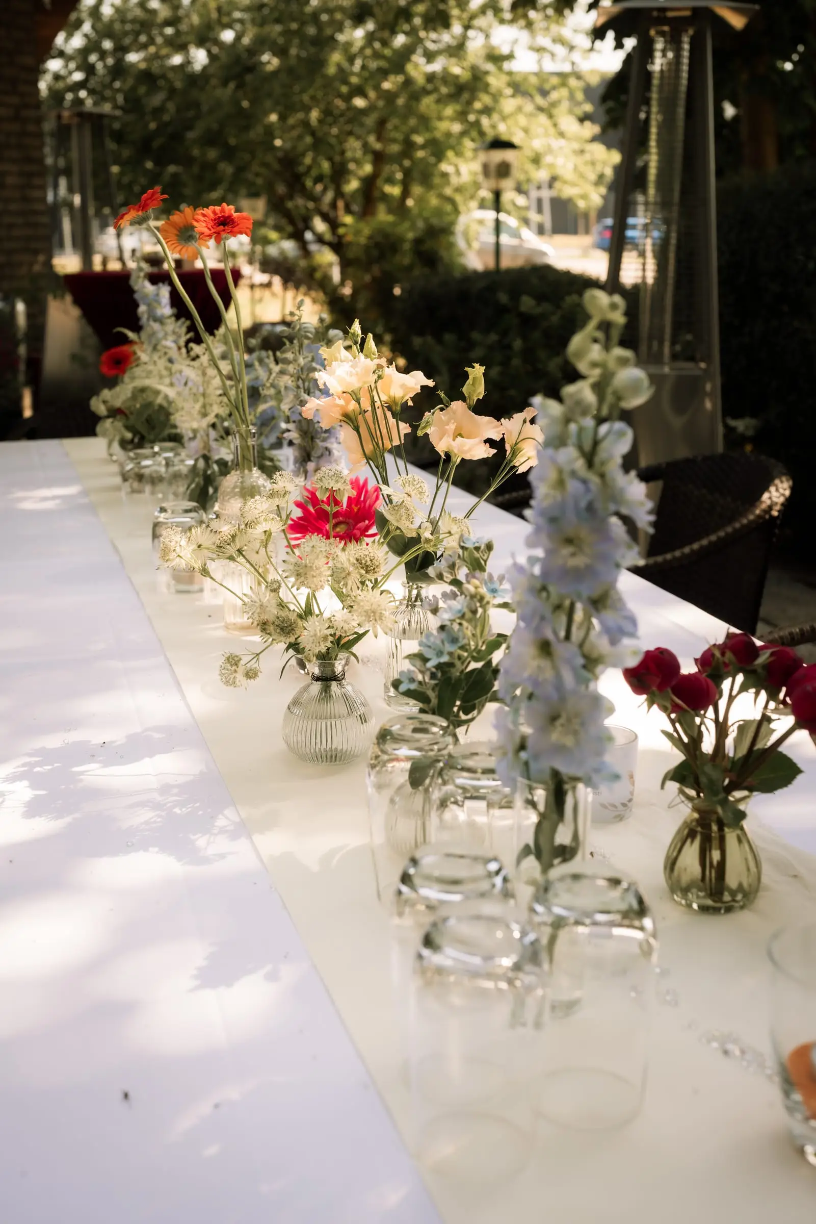 Lange weiße Hochzeitstafel im Freien unter Bäumen, dicht besetzt mit Blumen in kleinen Vasen