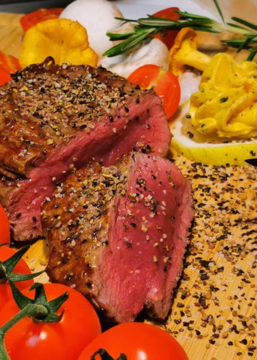 Aufgeschnittenes rosa Steak mit Pfefferkruste, Kräuterbutter, Pfifferlingen und Tomaten auf Holzbrett